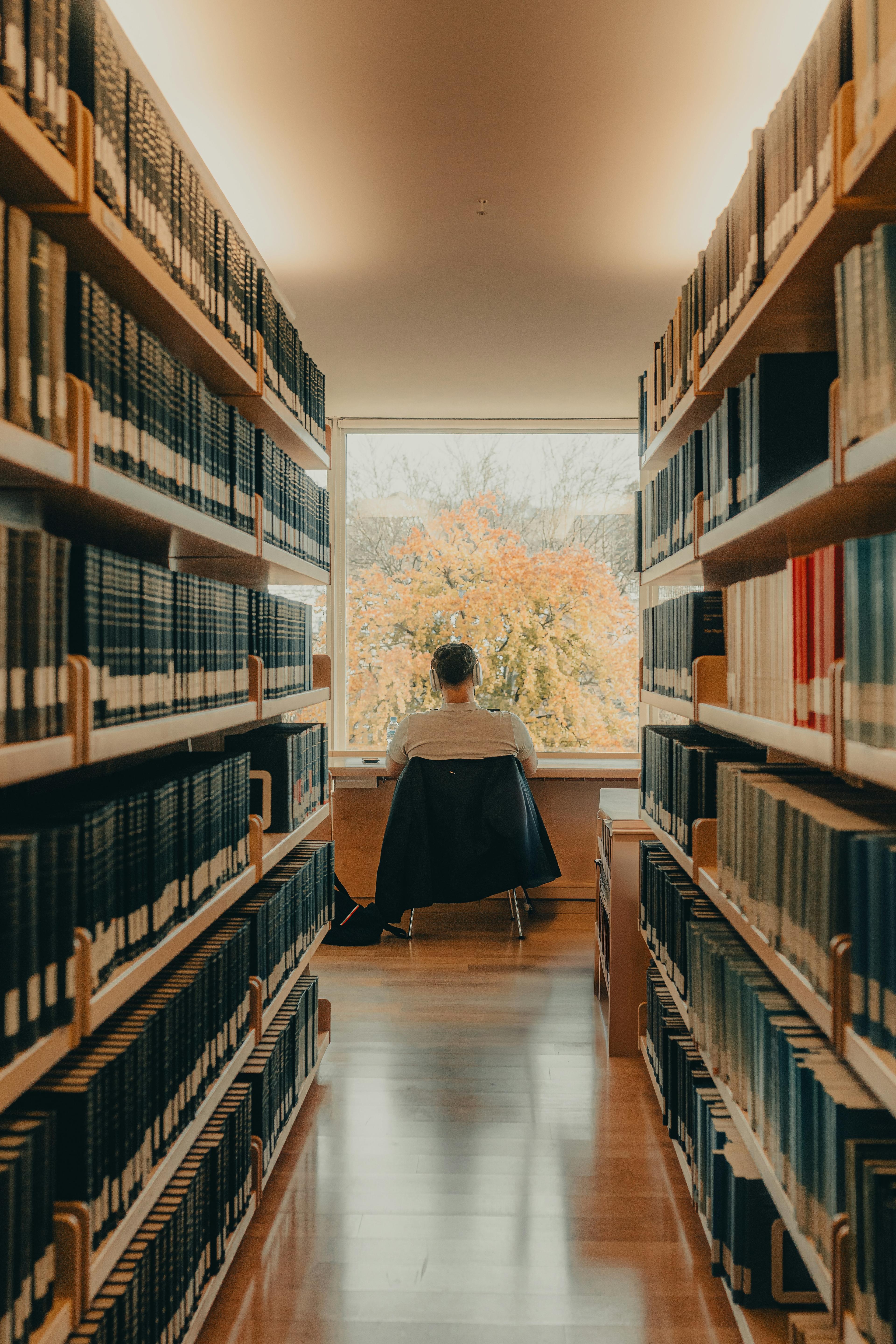 A reader by a window at the end of a long aisle of bookshelves
